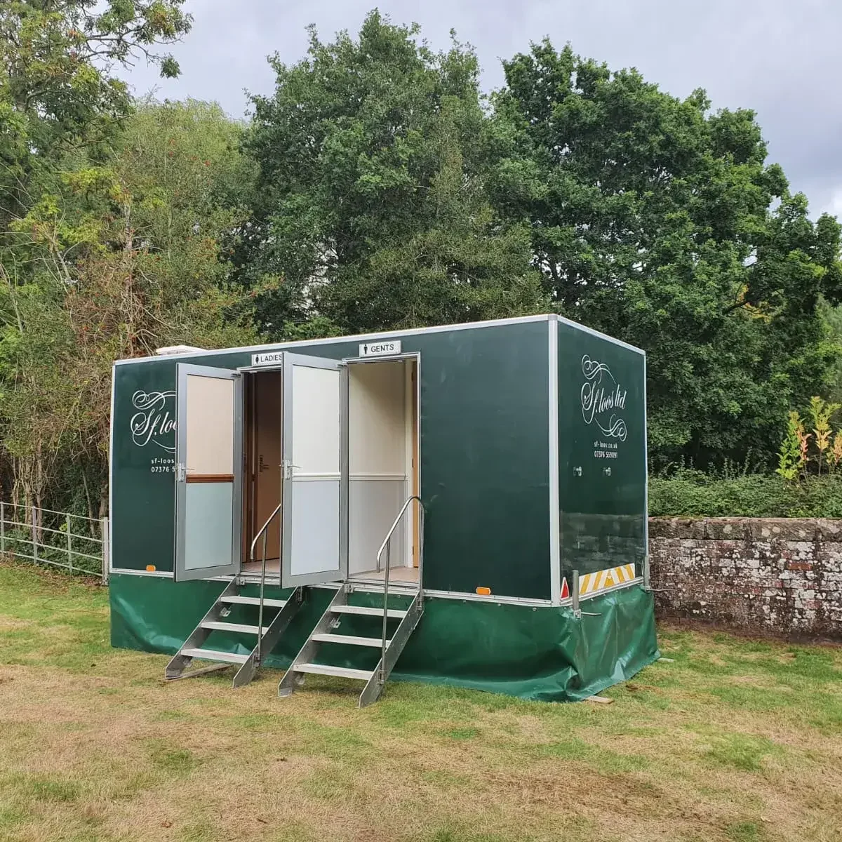 A portable restroom unit with two sections labeled "Ladies" and "Gents" is placed on a grassy area. Each section has its own door and steps leading up. The structure is backed by trees and a stone wall.